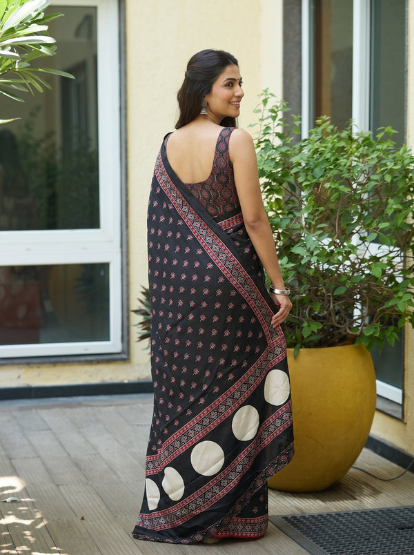 A woman wearing a black saree with red and gold patterns and white circular motifs, standing on a wooden deck next to a large yellow planter.