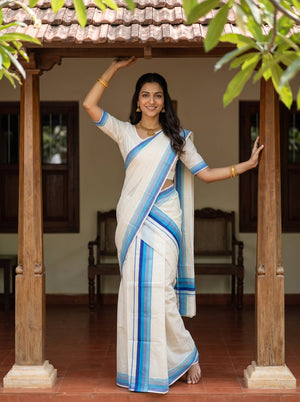 A woman wearing a white and blue saree stands under a wooden pavilion with tiled roofs, posing with her arms outstretched. The background features traditional wooden pillars, a tiled roof, and a bench.