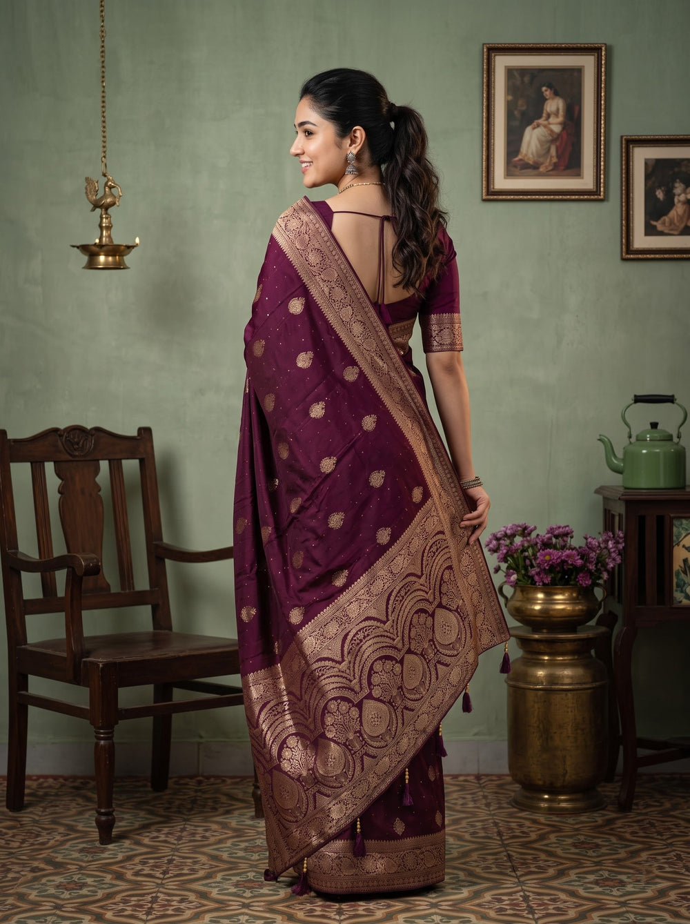 A woman modeling a deep purple silk saree with intricate gold and beige zari patterns and borders, standing in a rustic room with a green wall, wooden furniture, and a framed painting.