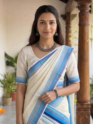 A woman wearing a cream-colored cotton saree with blue and purple striped borders, paired with a matching cream blouse, standing in a traditional indoor setting with wooden pillars.