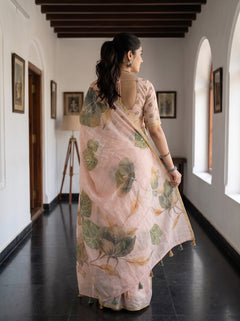A woman viewed from behind wearing a light pink saree with a green and beige floral print, walking down a hallway with white walls and dark wooden ceiling beams.