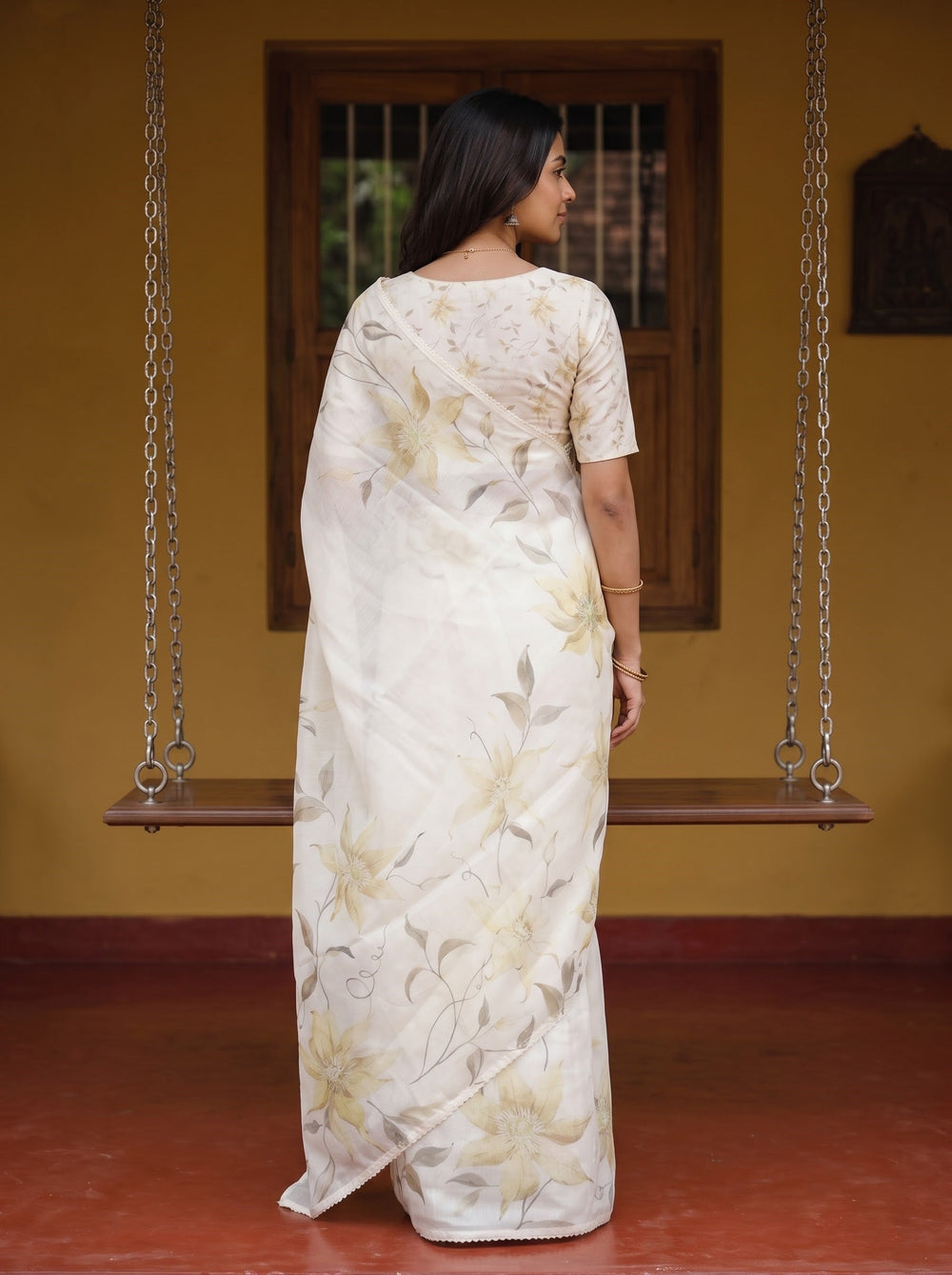 A woman wearing a cream-colored floral saree with a matching printed blouse stands in front of a wooden swing and a yellow wall.
