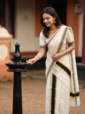 A woman wearing a cream-colored sari with black and gold stripes stands beside a black oil lamp, holding a lit match. The background shows a softly blurred traditional building interior.