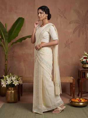 A woman models a cream-colored cotton saree with intricate silver embroidery along the border and pallu, paired with a matching embroidered blouse, in a styled studio setting with a terracotta wall and decorative plants.
