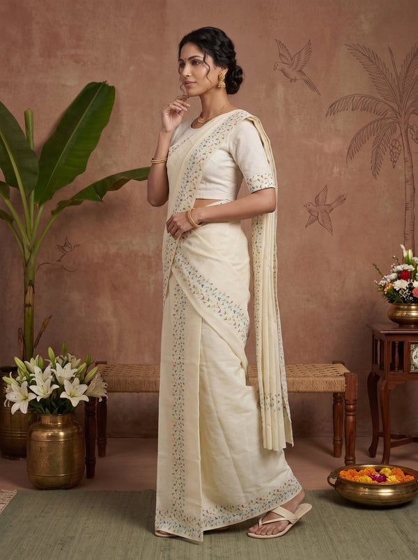 A woman models a cream-colored cotton saree with intricate silver embroidery along the border and pallu, paired with a matching embroidered blouse, in a styled studio setting with a terracotta wall and decorative plants.