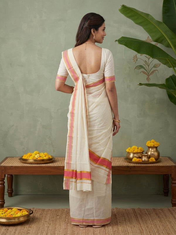 A woman stands with her back to the camera wearing a cream-colored cotton saree with pink and orange border stripes, paired with a matching cream blouse. The setting features a textured sage green wall, a wooden bench with yellow flower arrangements, and a large green plant leaf in the upper right.