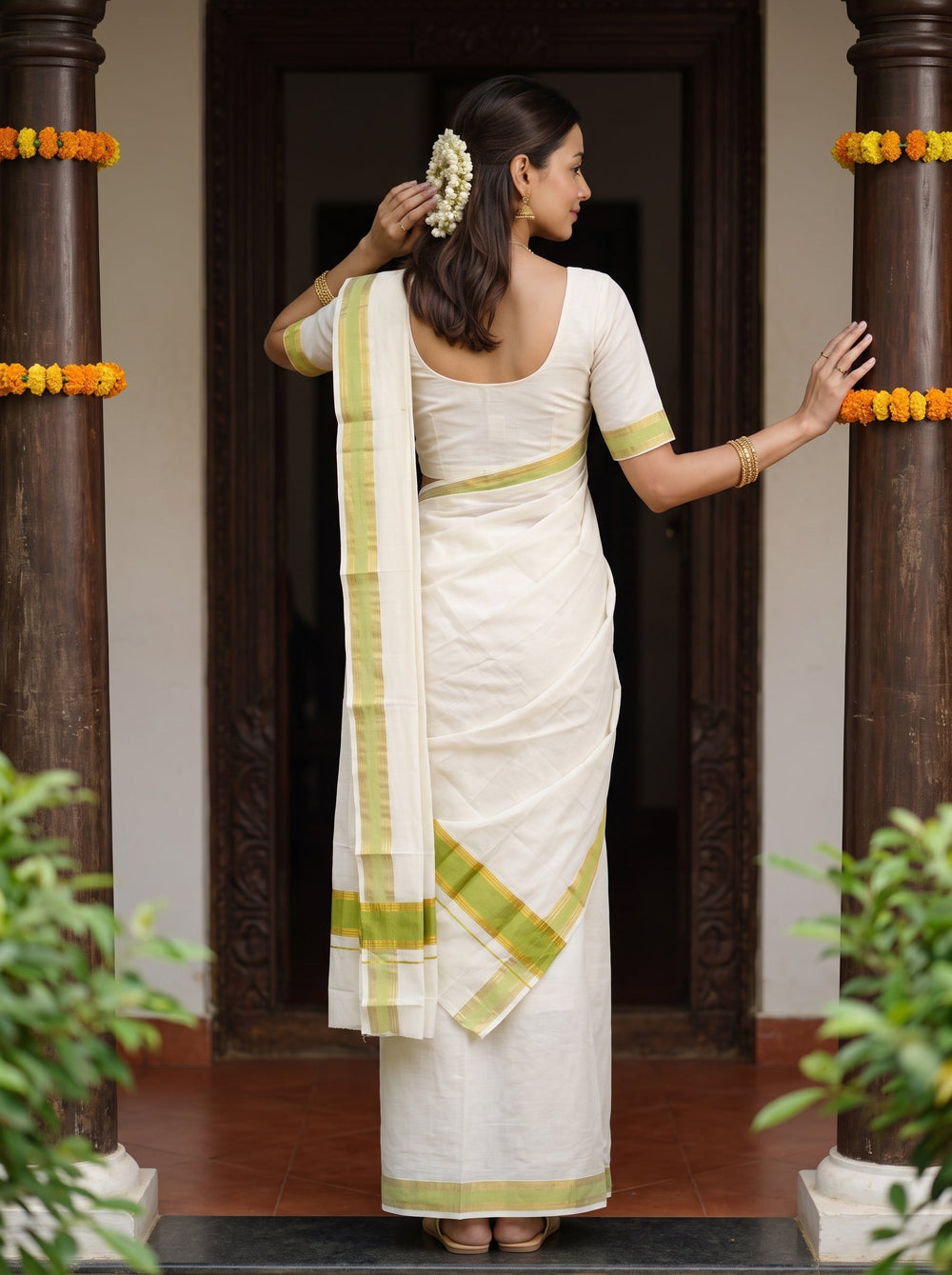 A woman wearing a white silk saree with lime green stripes stands in a doorway between two pillars decorated with yellow marigold garlands. She is holding a pink lotus flower.