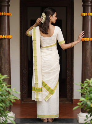 A woman wearing a white cotton saree with light green borders, standing with her back to the camera in a doorway decorated with dark pillars and yellow flower garlands.