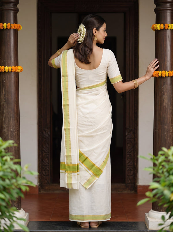 A woman wearing a white cotton saree with light green borders, standing with her back to the camera in a doorway decorated with dark pillars and yellow flower garlands.