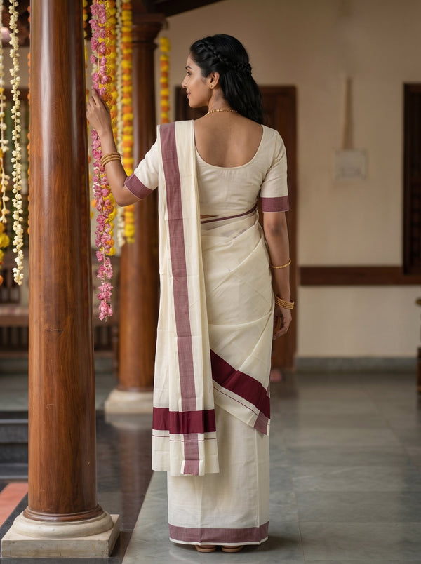 A woman wearing a cream-colored cotton saree with maroon borders, standing in a decorated indoor corridor with wooden pillars and hanging flower garlands.