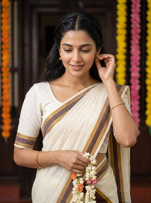 A woman wearing a cream-colored traditional sari with brown and gold stripes, holding a white flower garland. She is standing indoors in front of a dark wooden door with colorful flower garlands hanging vertically in the background.