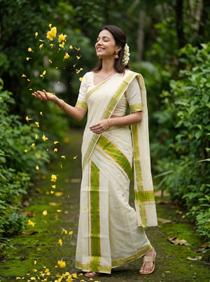 A woman wearing a cream-colored cotton saree with lime green stripes and border details, standing on a mossy forest path with yellow flower petals floating around her.