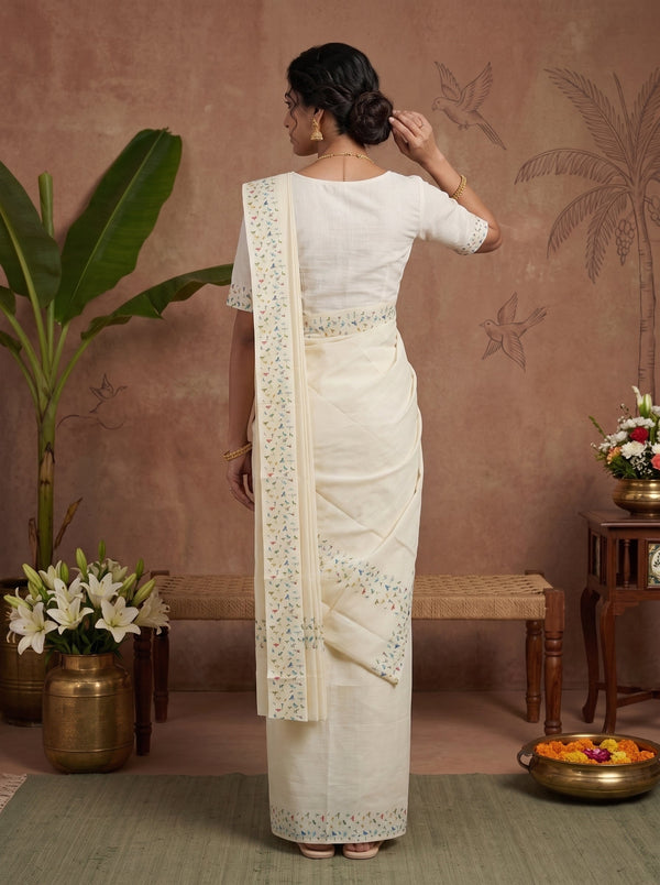 A woman stands with her back to the camera wearing a cream-colored cotton saree with a matching blouse. The saree features delicate floral embroidery in light blue and green along the border and pallu.