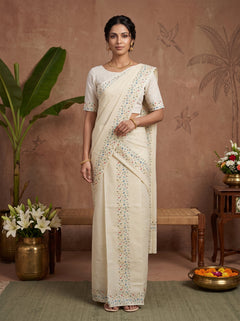 A woman stands indoors wearing a cream-colored saree with a matching short-sleeve blouse. The saree has delicate embroidered details along the border and drape, and the background features a textured terracotta wall with decorative plants and furniture.