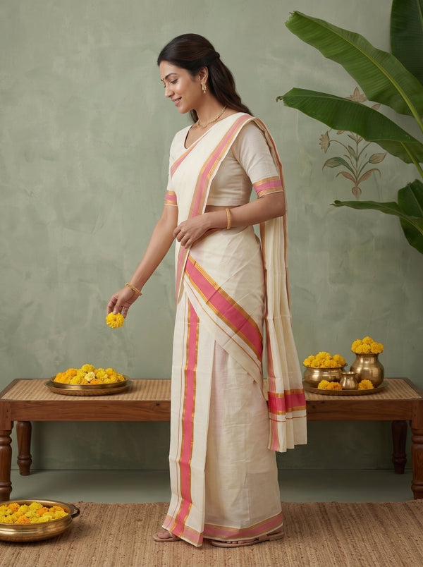 A woman wearing a cream-colored sari with pink and orange border stripes stands in a studio setting, holding yellow flowers near a wooden table. The background features a textured sage green wall with a painted floral motif and a large green plant leaf.