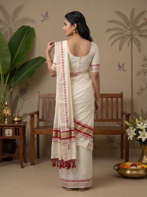 A woman stands with her back to the camera wearing a cream-colored Indian saree with red geometric border patterns and a matching cream blouse. The setting is a decorated room with a wooden bench, a potted plant, and floral arrangements.