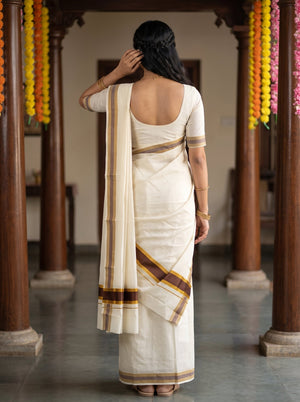 A woman viewed from behind wearing a cream-colored cotton saree with brown and gold stripes, standing in a traditional hall with wooden pillars and hanging flower garlands.