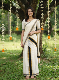 A woman wearing a cream-colored cotton saree with black and gold stripes, standing outdoors in front of a tree with hanging marigold garlands.
