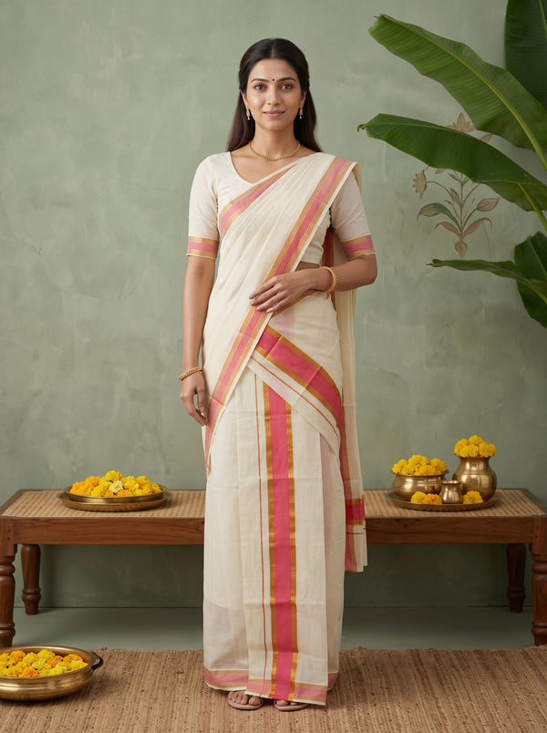 A woman stands wearing a cream-colored sari with pink and gold borders, paired with a matching cream blouse. The setting features a textured sage green wall, a wooden bench with yellow flowers, and a large green plant leaf in the background.