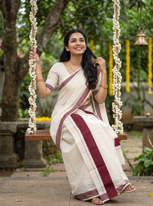 A woman wearing a cream-colored sari with maroon borders sits on a wooden swing decorated with white flower garlands in an outdoor garden setting.