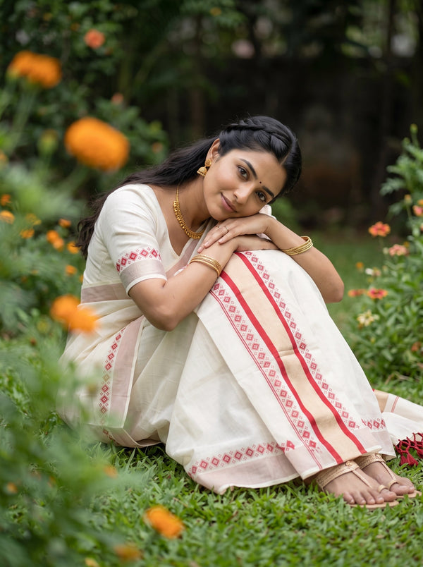 A woman wearing a cream-colored traditional Indian saree with red border patterns sits on green grass in a garden with orange flowers.