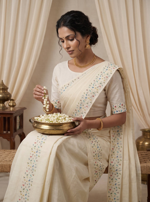 A woman seated indoors wearing a cream-colored saree with small multicolored floral embroidery along the border and pallu, holding a brass bowl filled with white flowers. The background features draped cream fabric curtains and warm wooden furniture.