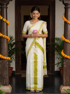 A woman wearing a white silk saree with lime green stripes stands in a doorway between two pillars decorated with yellow marigold garlands. She is holding a pink lotus flower.