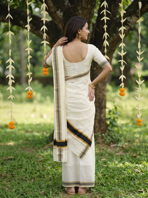 A woman wearing a cream-colored cotton saree with a black and gold border, standing outdoors in front of a tree with hanging marigold garlands.